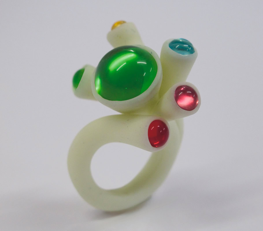 Bague blanche avec des pierres vertes, rouges et bleues sur un fond blanc.