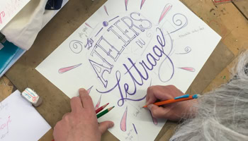 Personne faisant de la calligraphie sur une feuille de papier avec des crayons, avec les mots "ATELIERS de lettrage" visibles.