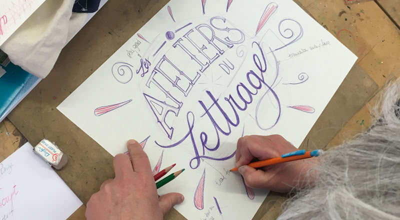 Personne faisant de la calligraphie sur une feuille de papier avec des crayons, avec les mots "ATELIERS de lettrage" visibles.