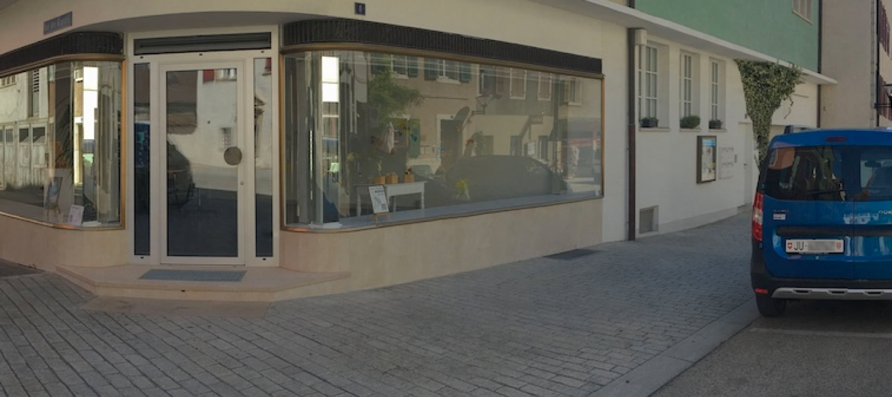 Rue pavée avec des bâtiments de chaque côté, une voiture bleue garée à droite et une vitrine d'angle à gauche.