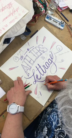 Personne faisant de la calligraphie sur une feuille de papier avec des crayons, avec les mots "ATELIERS de lettrage" visibles.