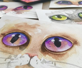 Peintures aquarelles de visages de chats avec de grands yeux colorés, principalement violets et verts, disposées sur une surface.