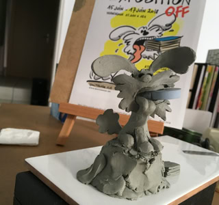 Sculpture en argile d'un personnage avec de grandes oreilles sur une table, avec une affiche colorée en arrière-plan.