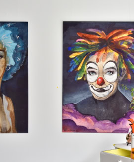 Deux peintures accrochées au mur, l'une représentant une personne avec des cheveux bleus et l'autre un clown avec des cheveux multicolores. Sur une table en dessous, des objets décoratifs et une plante.