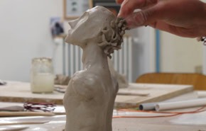 Une main sculpte une tête en argile sur une table, avec des outils et des pots en arrière-plan.
