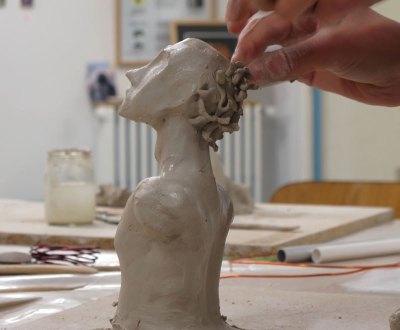 Une main sculpte une tête en argile sur une table, avec des outils et des pots en arrière-plan.