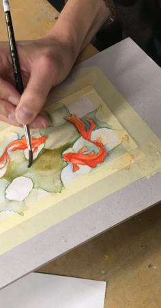 Personne peignant une aquarelle sur une feuille de papier fixée avec du ruban adhésif, pinceaux à proximité.