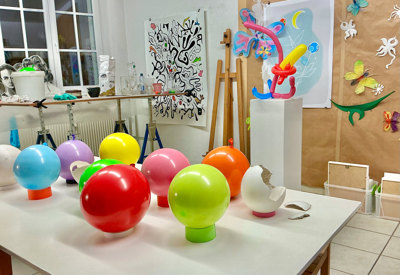 Salle d'art avec des ballons colorés sur une table, des œuvres d'art accrochées aux murs, et divers objets artistiques exposés.