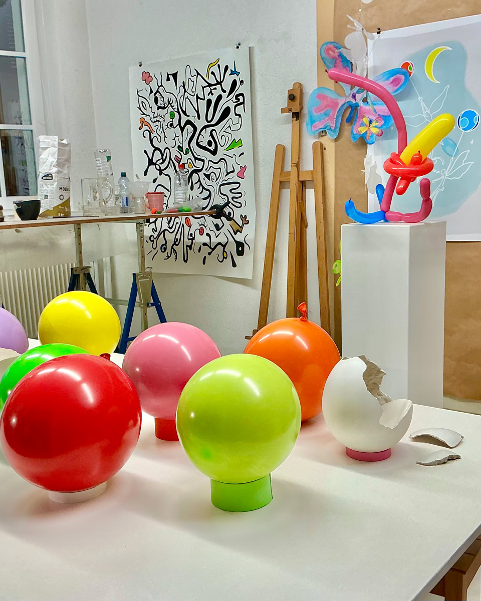 Salle d'art avec des ballons colorés sur une table, des œuvres d'art accrochées aux murs, et divers objets artistiques exposés.