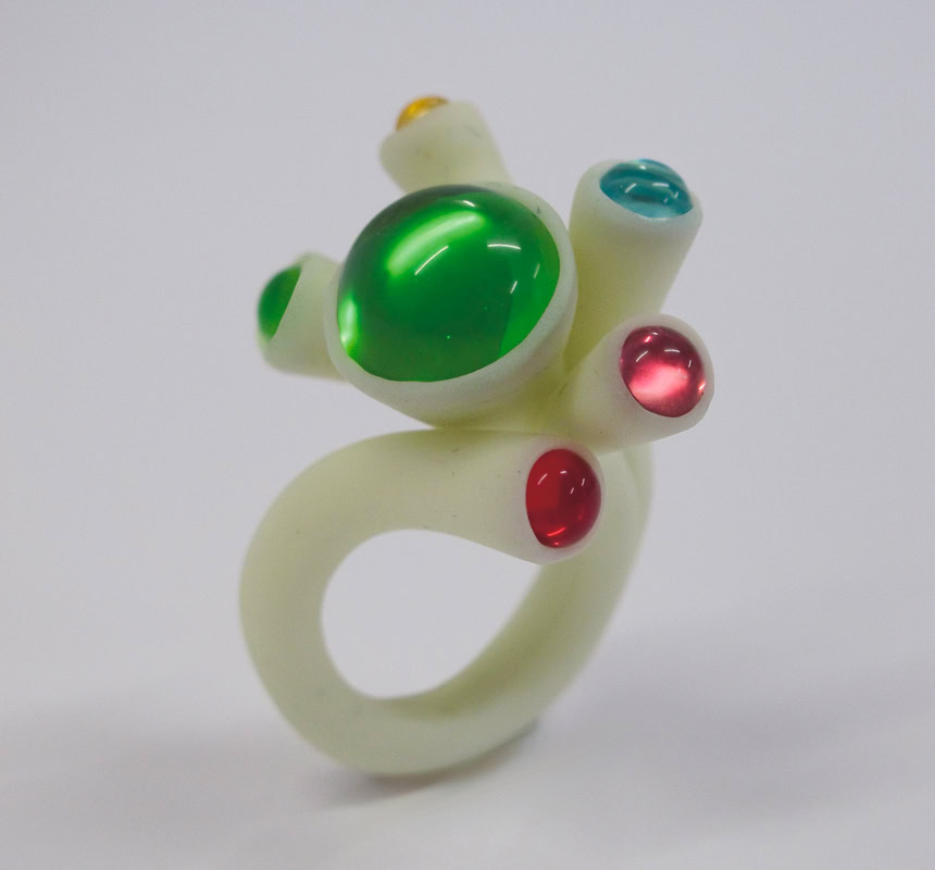 Bague blanche avec des pierres vertes, rouges et bleues sur un fond blanc.