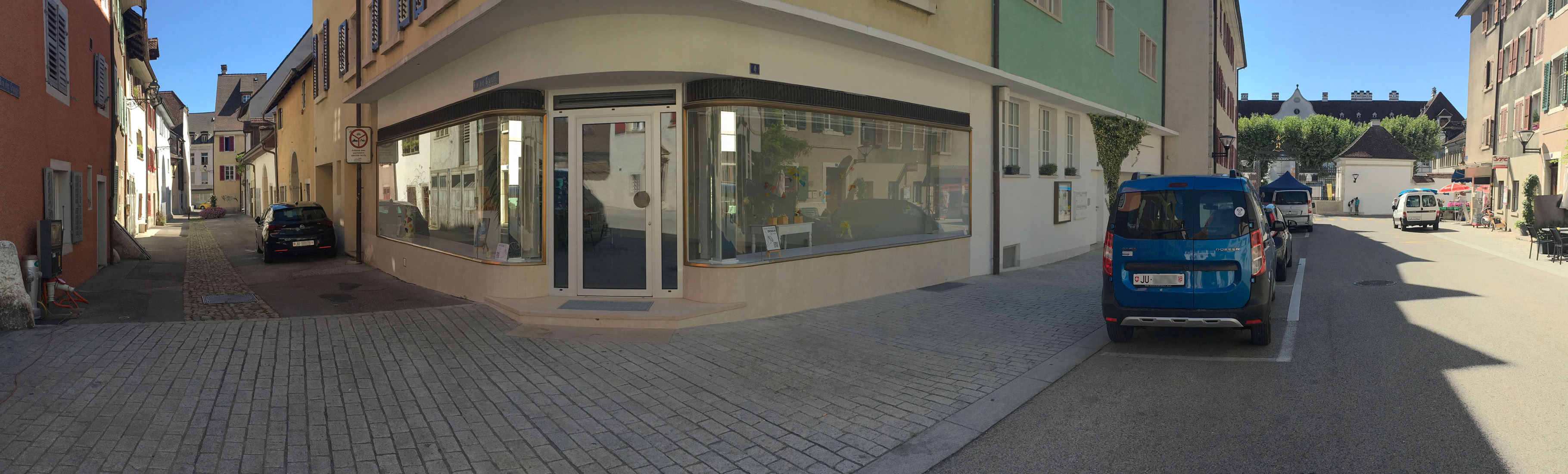 Rue pavée avec des bâtiments de chaque côté, une voiture bleue garée à droite et une vitrine d'angle à gauche.