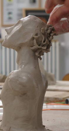 Une main sculpte une tête en argile sur une table, avec des outils et des pots en arrière-plan.