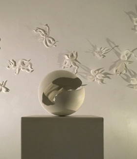 Sculpture en forme de sphère sur un socle, entourée de papillons blancs en relief sur un mur.