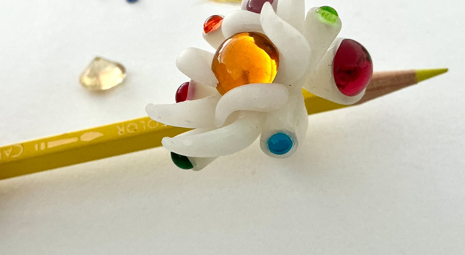 Un crayon rose traversant une bague en résine blanc avec des embouts colorés, entouré de crayons de couleur sur une table.
