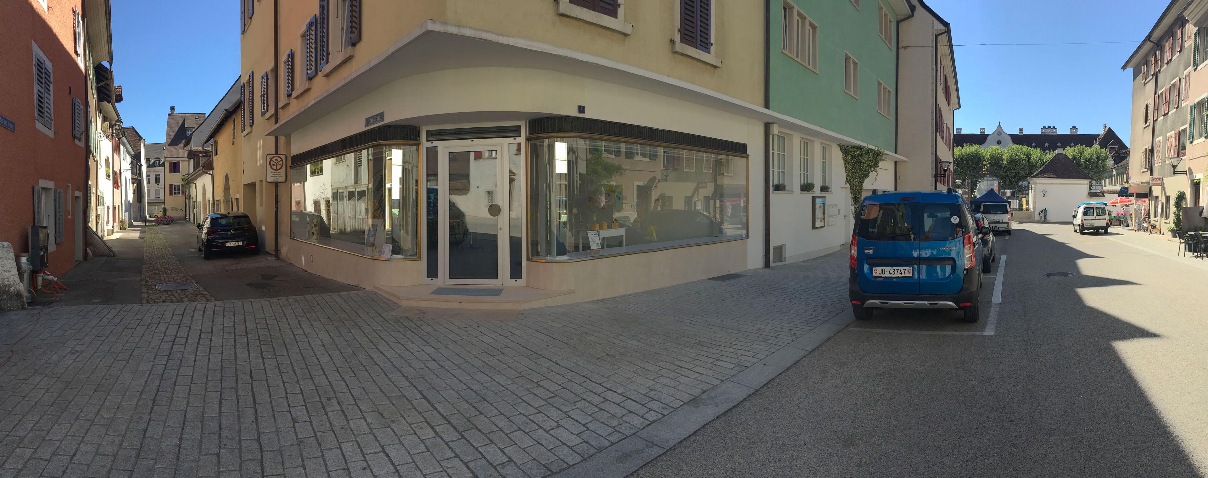 Rue pavée avec des bâtiments de chaque côté, une voiture bleue garée à droite et une vitrine d'angle à gauche.