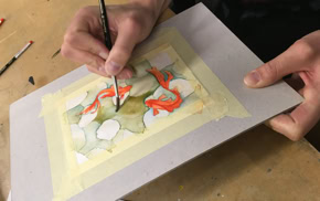 Personne peignant une aquarelle sur une feuille de papier fixée avec du ruban adhésif, pinceaux à proximité.