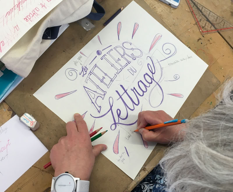 Personne faisant de la calligraphie sur une feuille de papier avec des crayons, avec les mots "ATELIERS de lettrage" visibles.