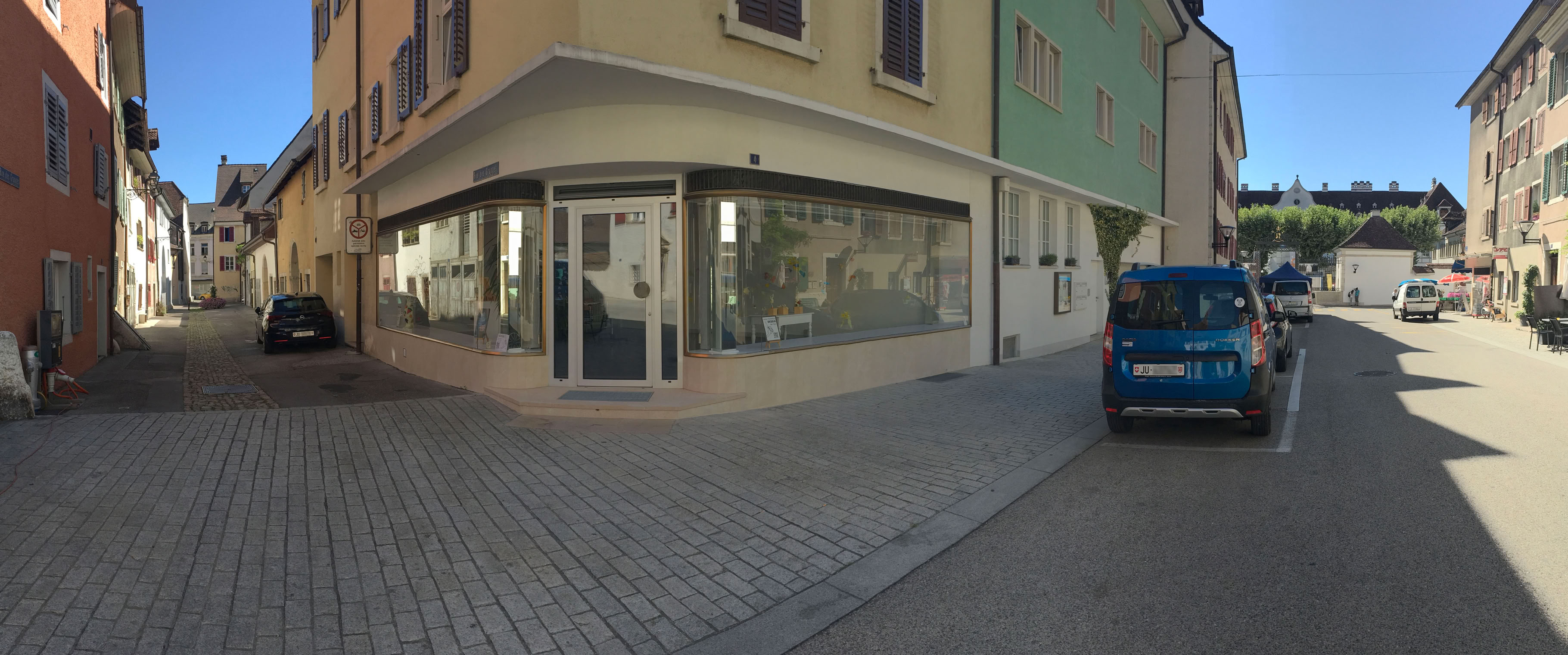 Rue pavée avec des bâtiments de chaque côté, une voiture bleue garée à droite et une vitrine d'angle à gauche.