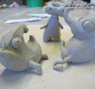 Trois petites sculptures en argile représentant des créatures stylisées avec de grands yeux et des formes arrondies, posées sur une surface de travail.