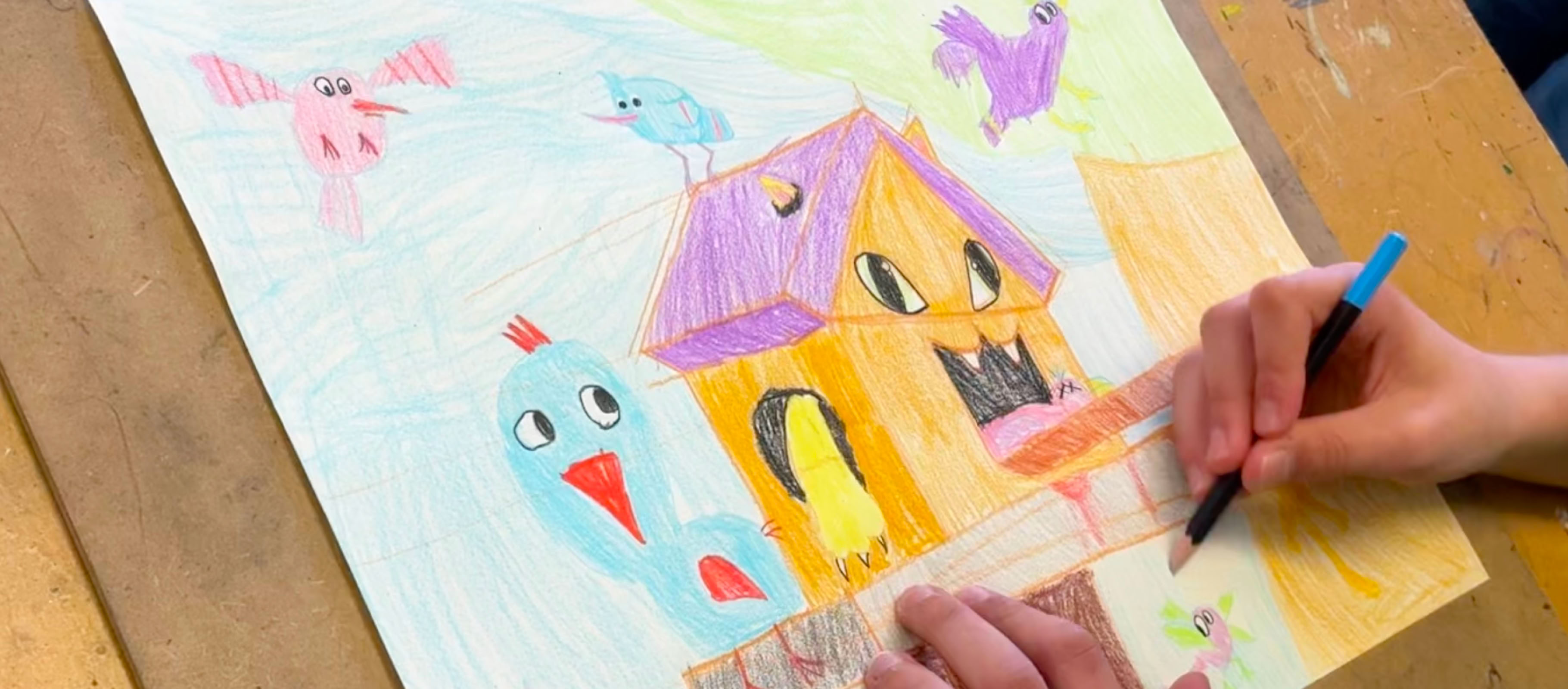 Dessin coloré d'une maison avec des oiseaux autour, une main tenant un crayon à côté.