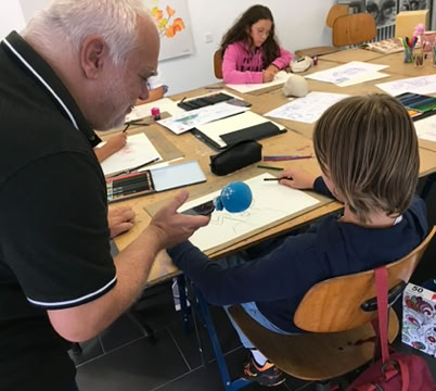 Un homme tend un microphone à un enfant assis à une table avec d'autres enfants, entourés de matériel de dessin.