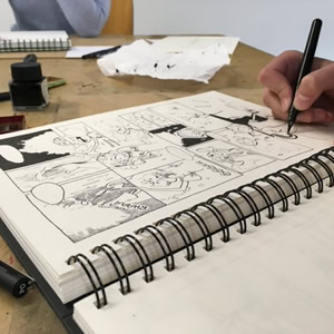 Personne dessinant dans un carnet à spirale avec un stylo, encre et feuilles de papier sur la table.