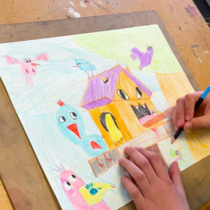 Une personne dessine une maison colorée avec des oiseaux sur une feuille de papier.