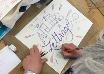 Personne faisant de la calligraphie sur une feuille de papier avec des crayons, avec les mots "ATELIERS de lettrage" visibles.